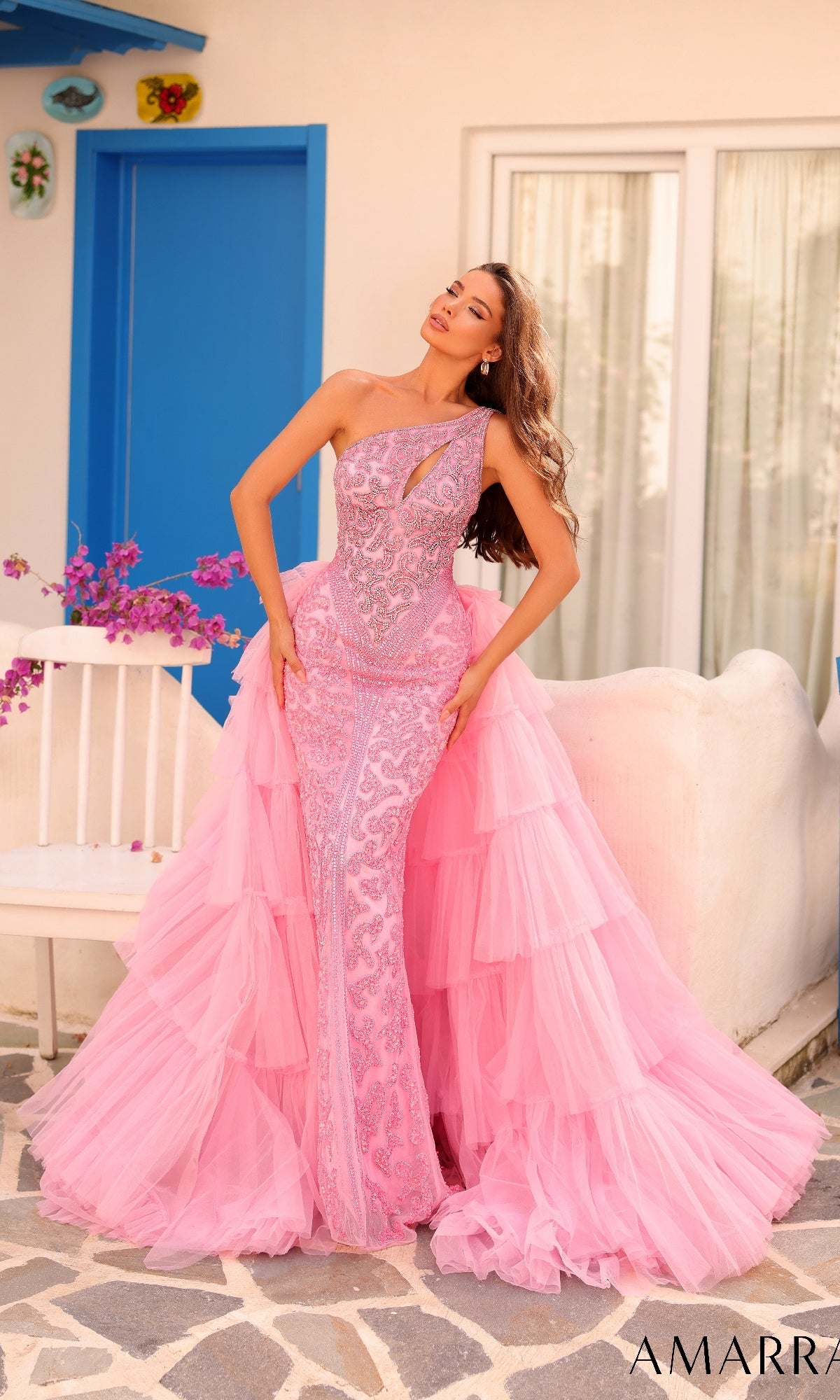 A woman poses outdoors in the Long Formal Dress 88147 by Amarra, a light pink one-shoulder gown with lace details and a ruffle tiered overskirt. A white flower-adorned bench and blue door complete the elegant scene.